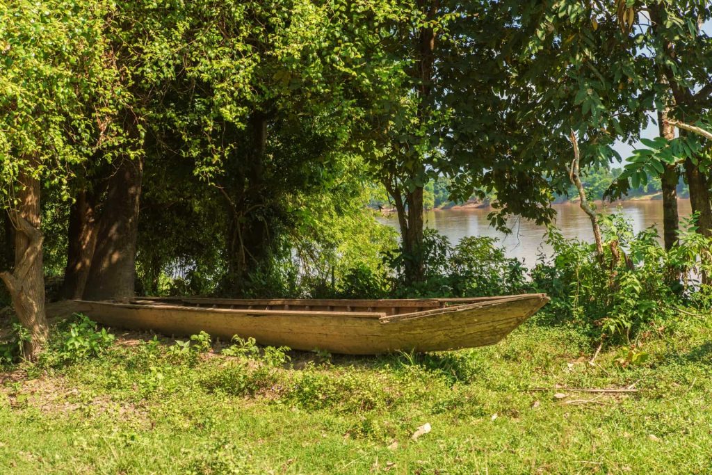A wooden boat rests on the grass beneath riverside trees.